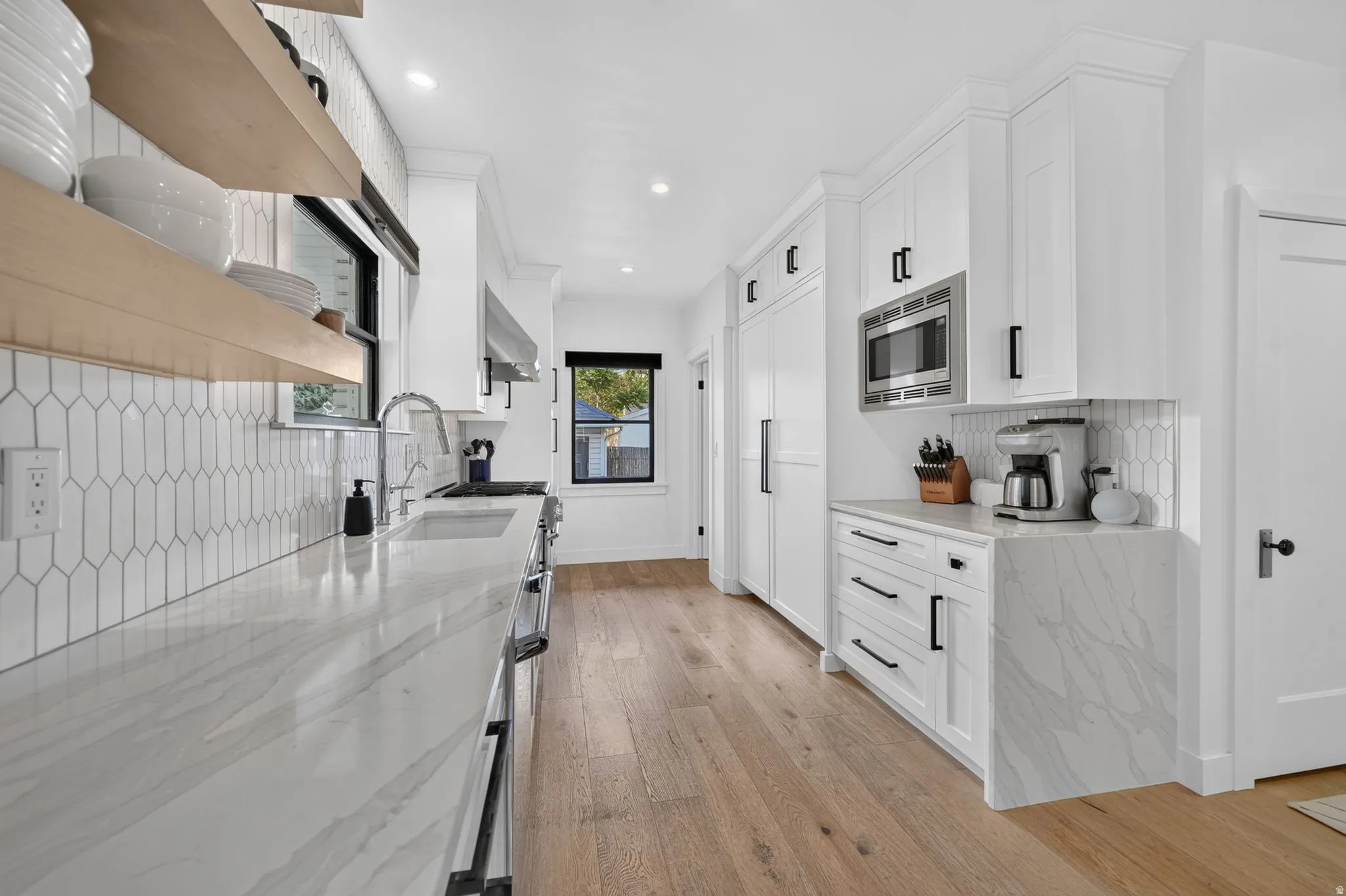 Stunning modern kitchen featuring white cabinetry, counter to ceiling tile backsplash, wood accent floating shelves, stainless steel appliances, cabinet-front refrigerator & freezer, and waterfall quartz countertops.
