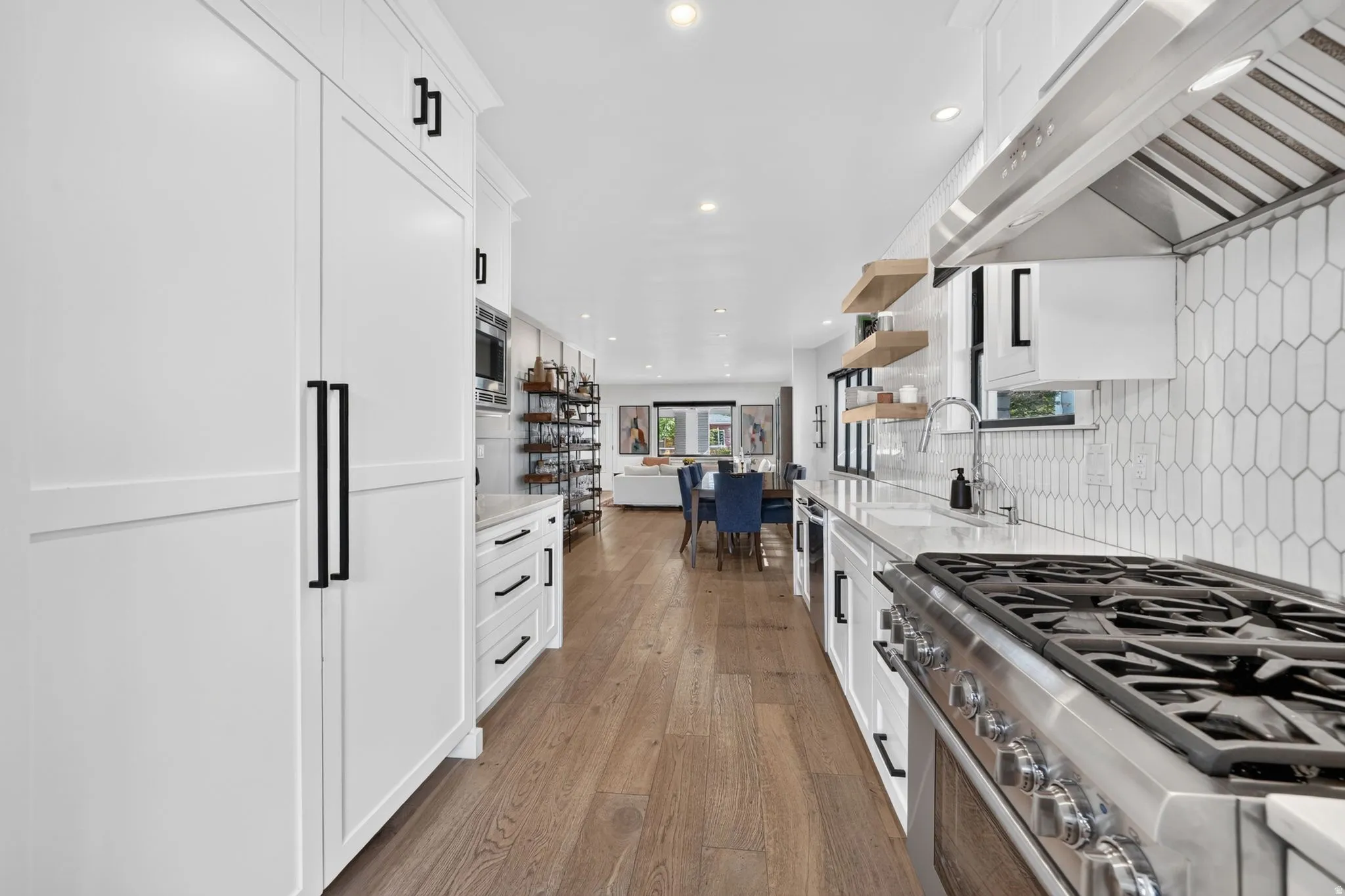 Gourmet kitchen featuring a stainless steel gas range with a professional-grade hood, tile backsplash, wood accent floating shelves, quartz countertops, and cabinet-front refrigerator & freezer