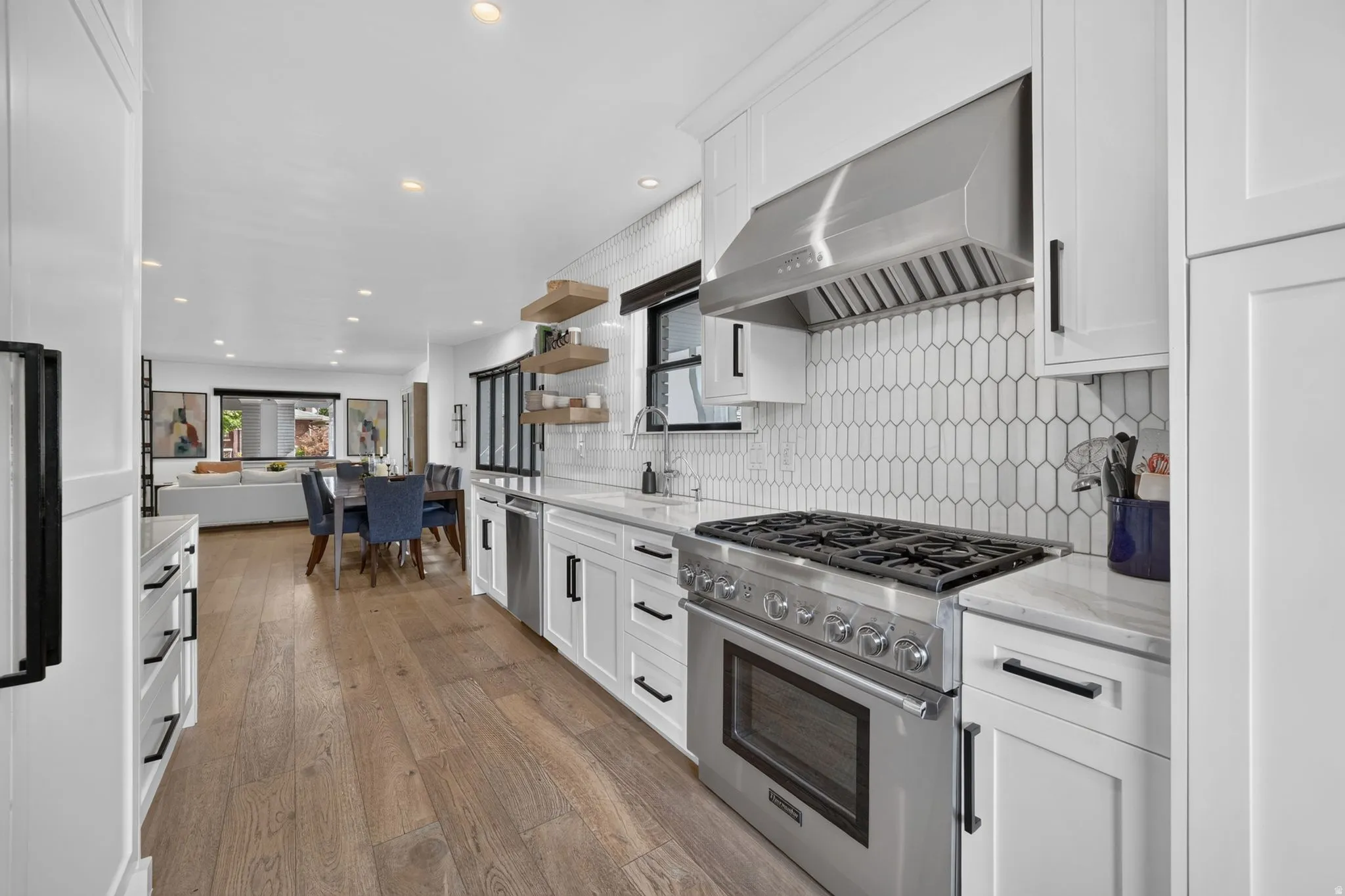 Gourmet kitchen featuring a stainless steel gas range with a professional-grade hood, tile backsplash, wood accent floating shelves, quartz countertops, and cabinet-front refrigerator & freezer