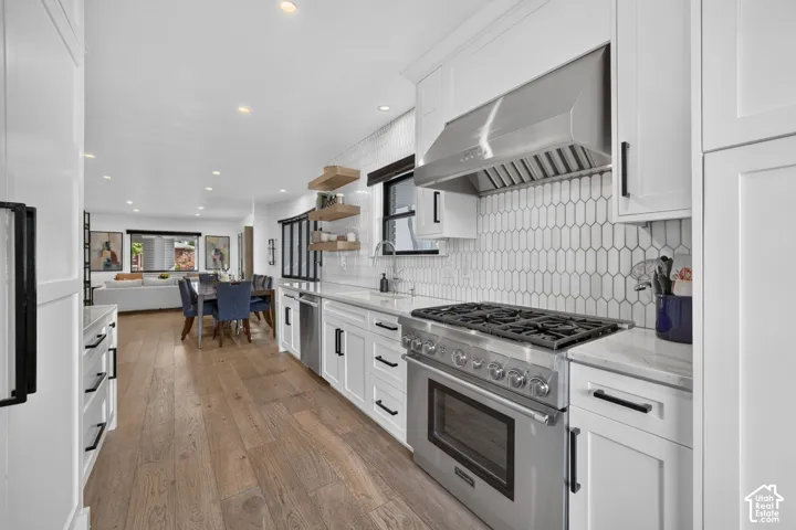 Gourmet kitchen featuring a stainless steel gas range with a professional-grade hood, tile backsplash, wood accent floating shelves, quartz countertops, and cabinet-front refrigerator & freezer