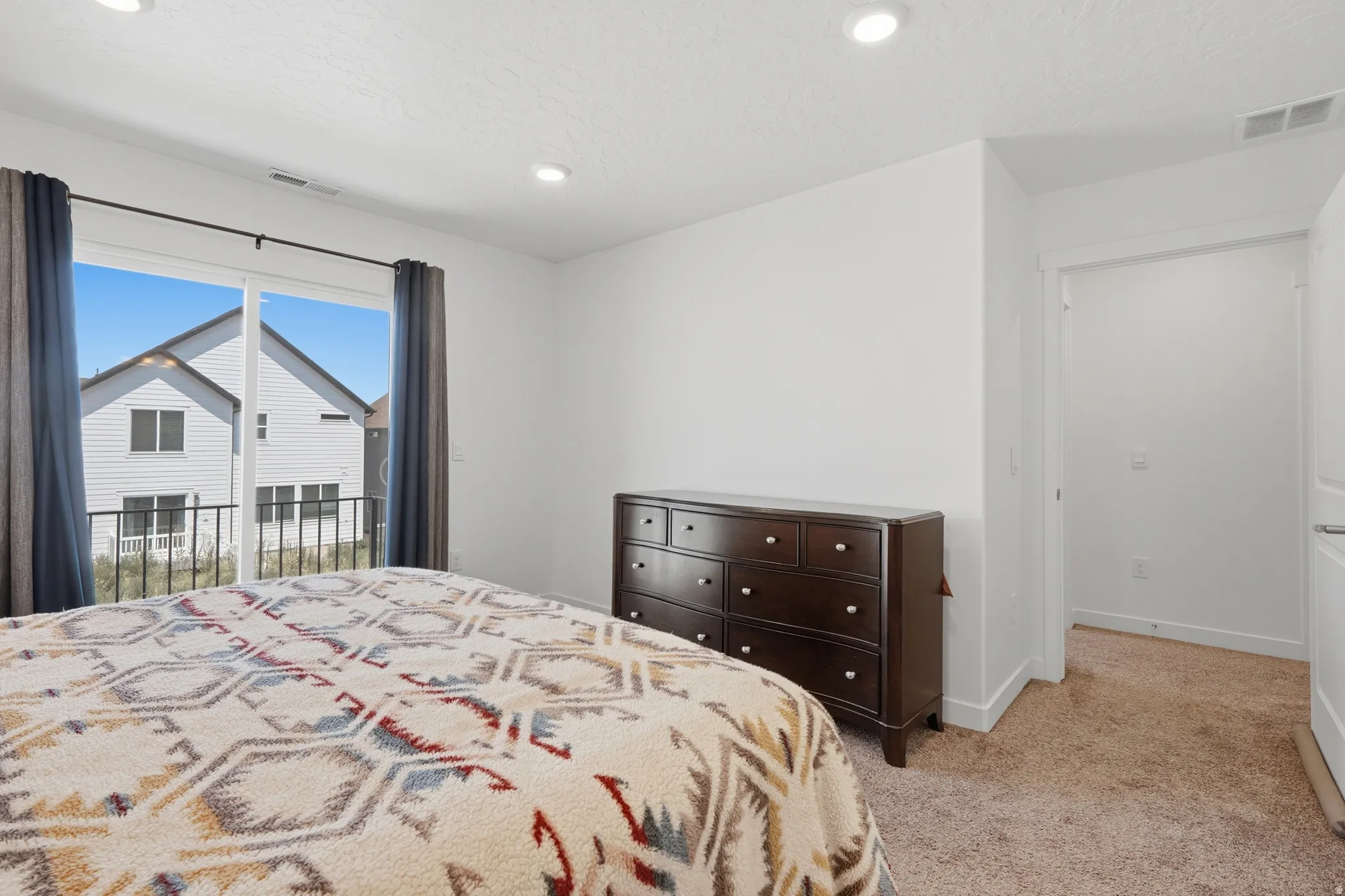 Bright bedroom featuring a sliding glass door to a balcony with black metal railing, recessed lighting, and neutral carpeting