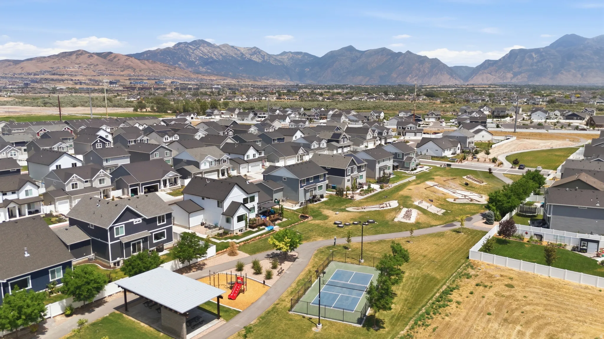 Residential development featuring a community park, covered picnic area, pickleball court, and playground equipment, all set against a prominent mountain backdrop
