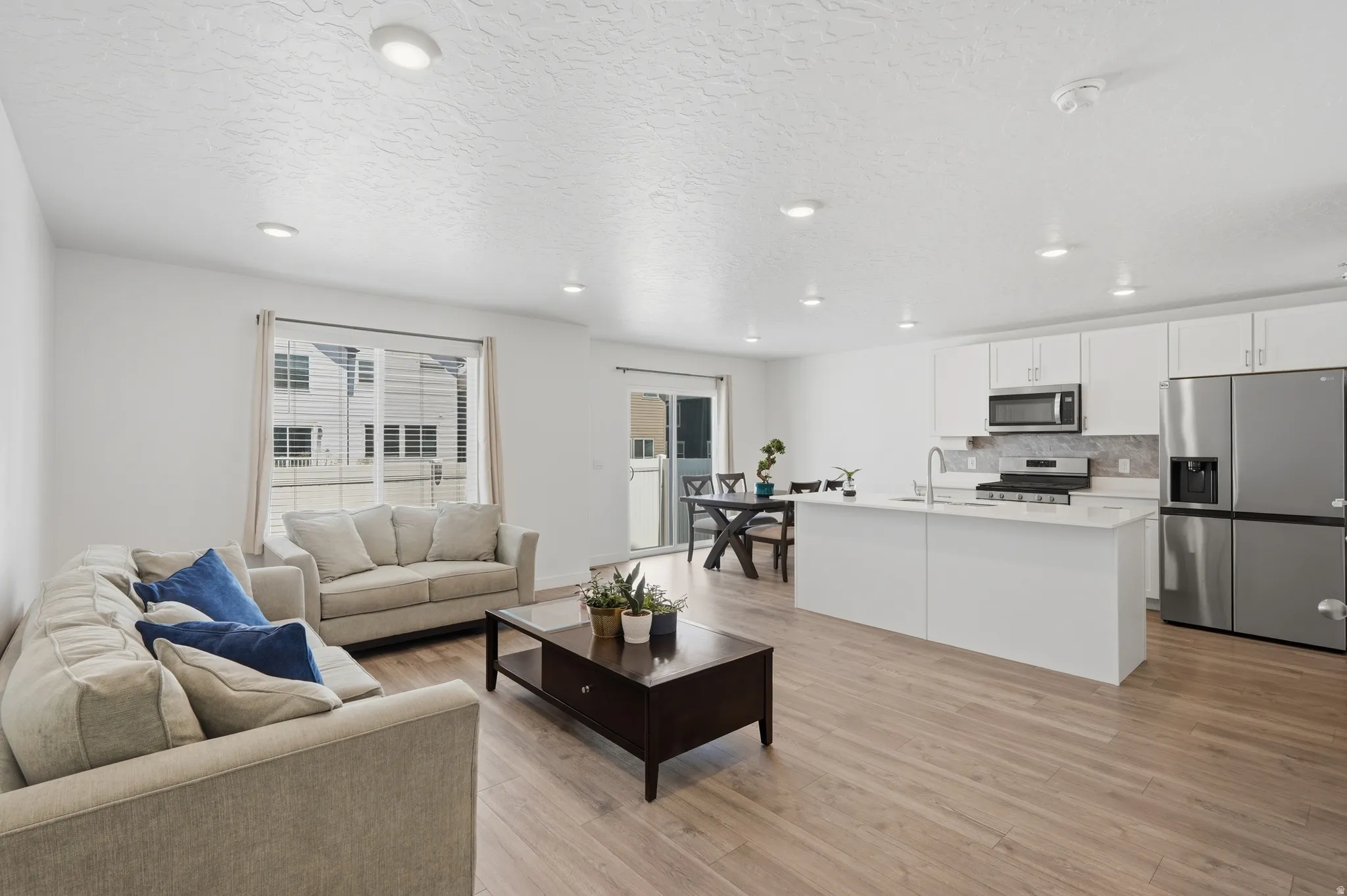 Open-concept living area featuring wood-finish flooring, recessed lighting, a kitchen island with sink, white cabinetry, and stainless steel appliances