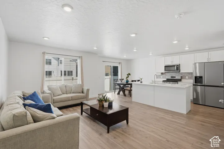 Open-concept living area featuring wood-finish flooring, recessed lighting, a kitchen island with sink, white cabinetry, and stainless steel appliances