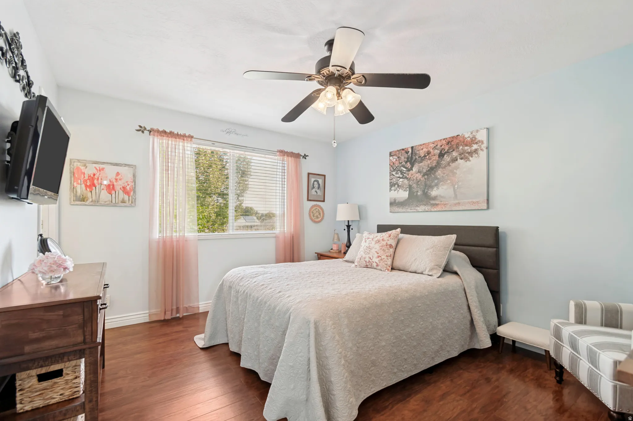 Wood-finish flooring throughout, featuring a ceiling fan with integrated lighting, window with blinds, and a built-in wall-mounted television bracket
