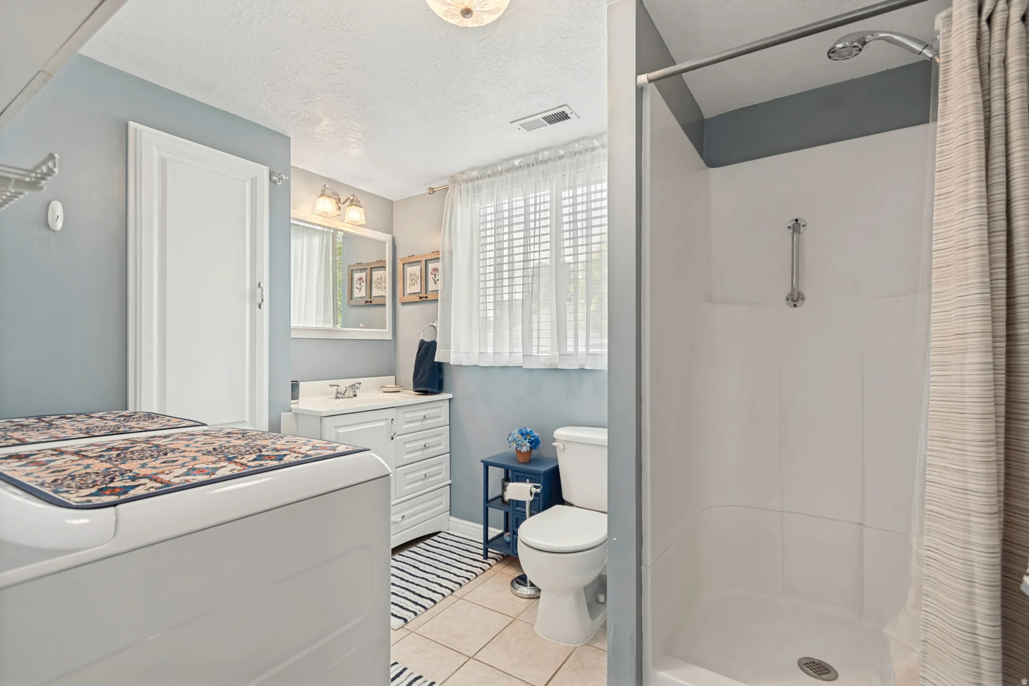 Bathroom/Laundry Room featuring a white vanity with integrated sink, window with privacy blinds, walk-in shower, and tile flooring