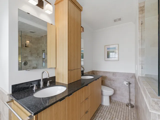 Bathroom featuring wood-finish cabinetry, black stone countertops, dual undermount sinks, and a tiled shower with glass enclosure