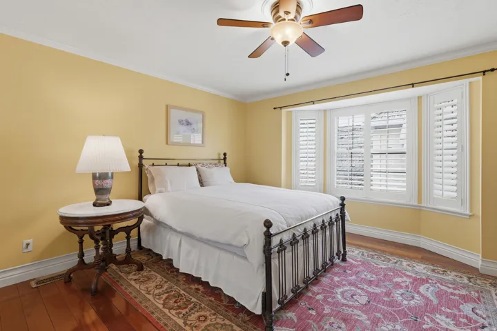 Bedroom featuring a multi-pane bay window with plantation shutters, crown molding, wood-finish flooring, and a ceiling fan with integrated lighting