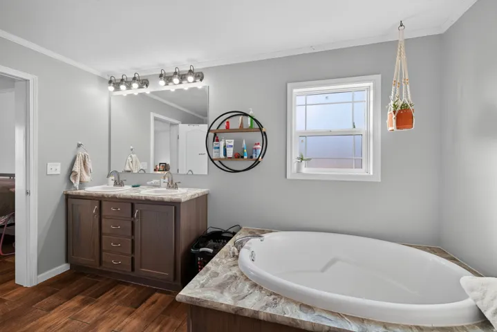 Built-in soaking tub with a patterned surround, double vanity with wood-finish cabinetry, dual sinks, and wood-finish flooring