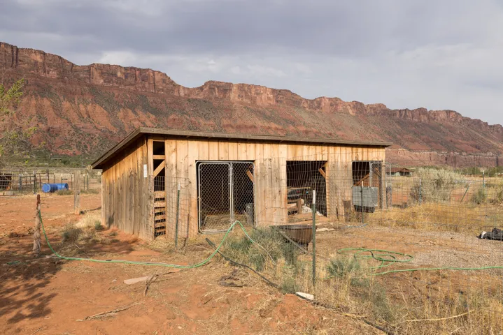 Wood-sided outbuilding with multiple chain-link enclosures, situated on a natural landscape with red rock formations in the background