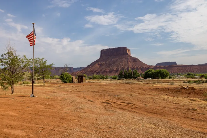 Expansive desert landscape featuring a clear sky, a prominent red rock mesa in the background, and scattered green trees