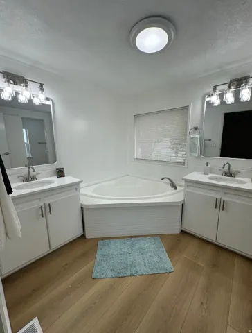 Bathroom featuring a corner soaking tub, dual white vanities with undermount sinks, wood-finish flooring, and a window with blinds
