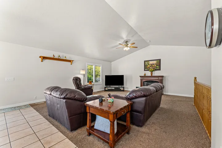 Carpeted living area featuring a vaulted ceiling with a ceiling fan, a fireplace with a wood mantle, a window with louvered shutters, and a tile entryway
