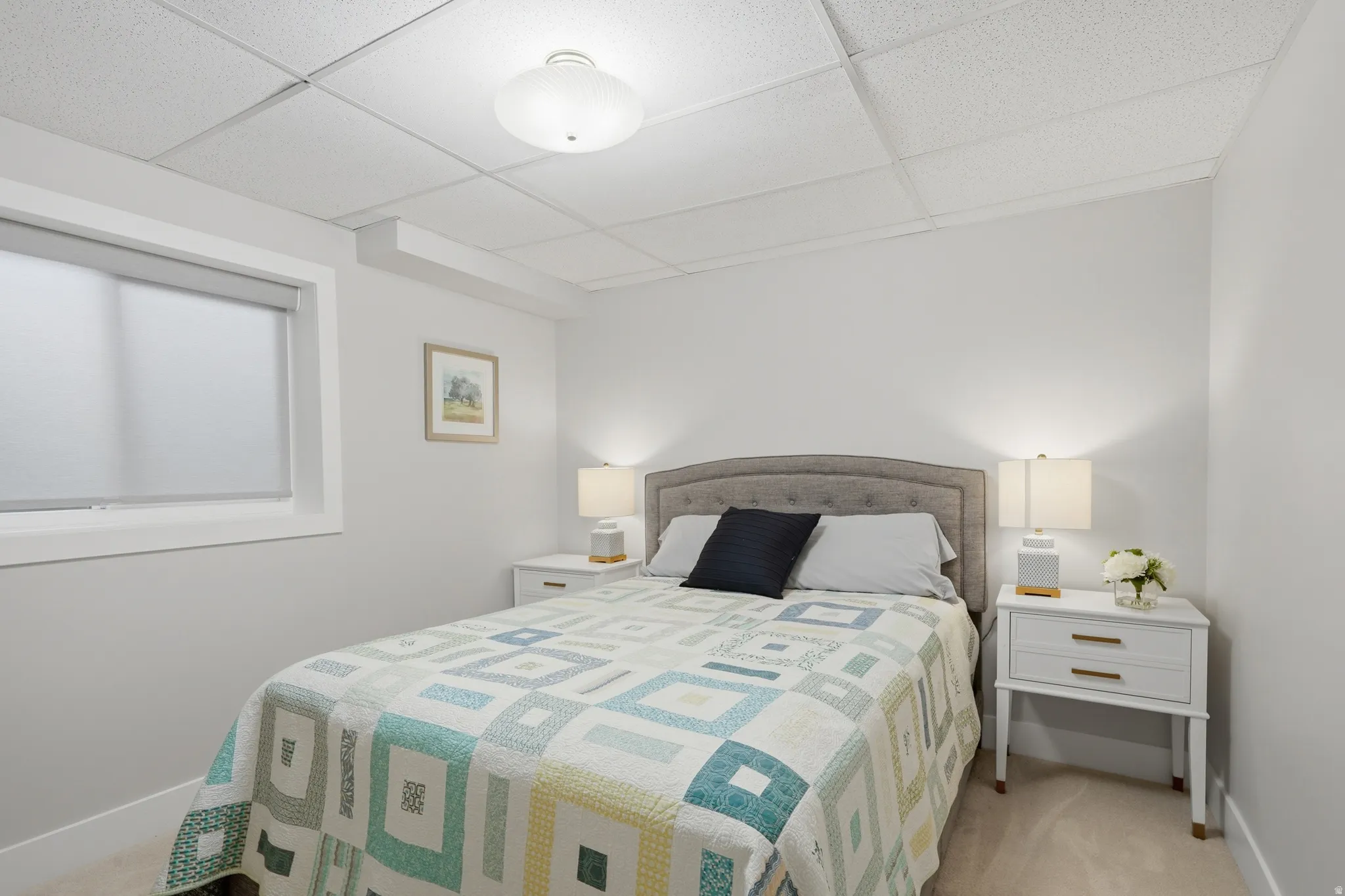 Bedroom featuring light-colored carpet flooring, a drop ceiling, and a single window with a roller shade