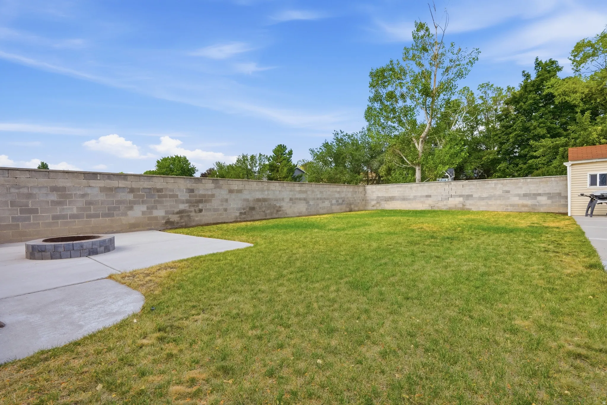 Spacious backyard featuring a concrete block perimeter wall, a dedicated concrete patio area, a built-in stone fire pit, and a separate structure with a shingled roof