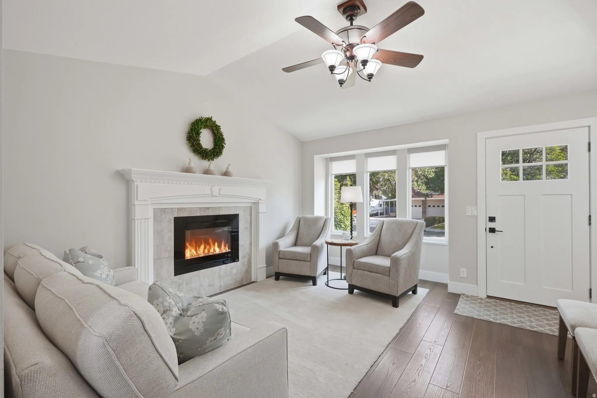 Living area featuring an electric fireplace with a white mantel and tile surround, vaulted ceiling, wood-finish flooring, and a white front door with upper lites