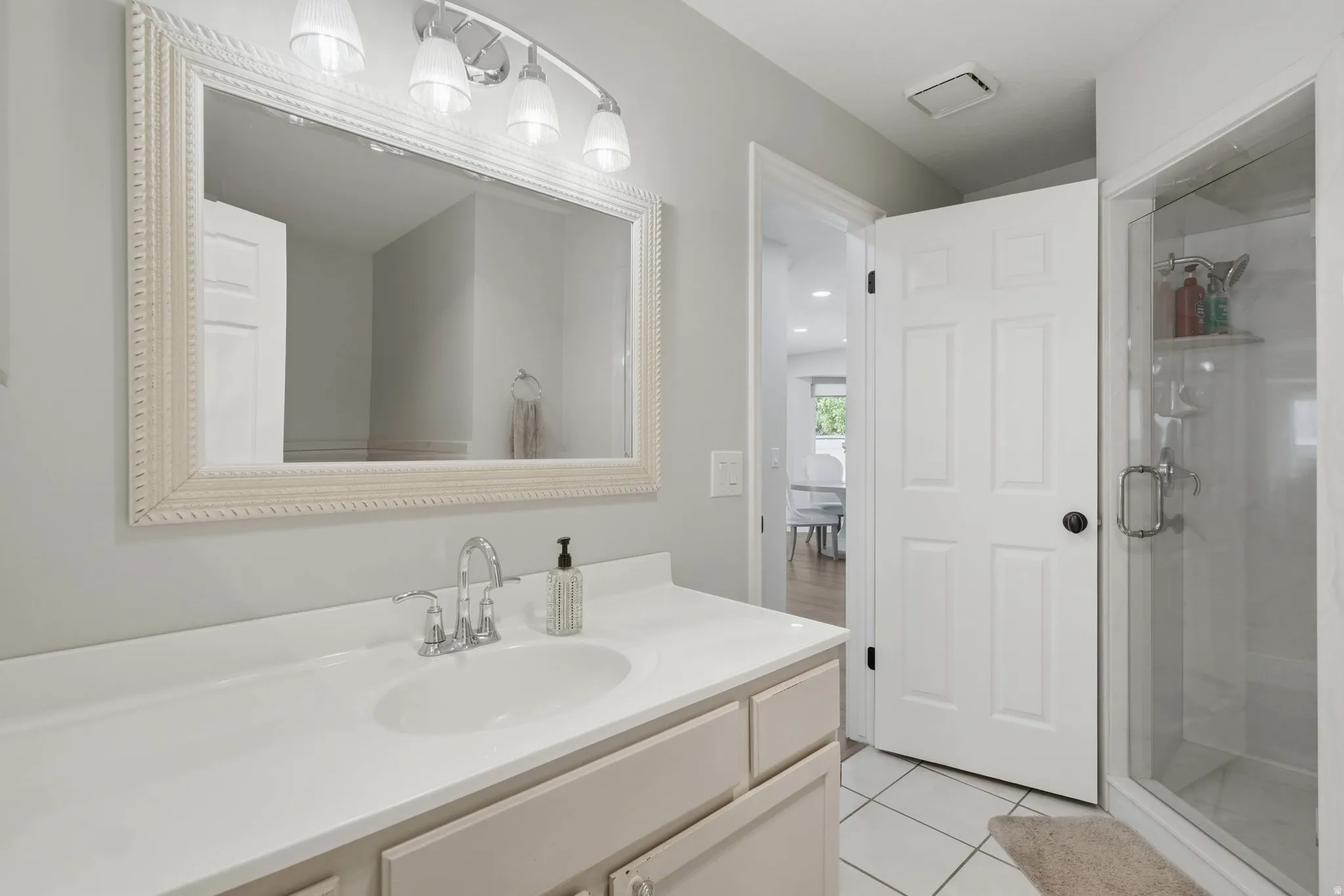 Bathroom featuring a single vanity with an integrated sink and chrome faucet, a large framed mirror, a four-light vanity fixture, a glass-enclosed shower, and white tile flooring