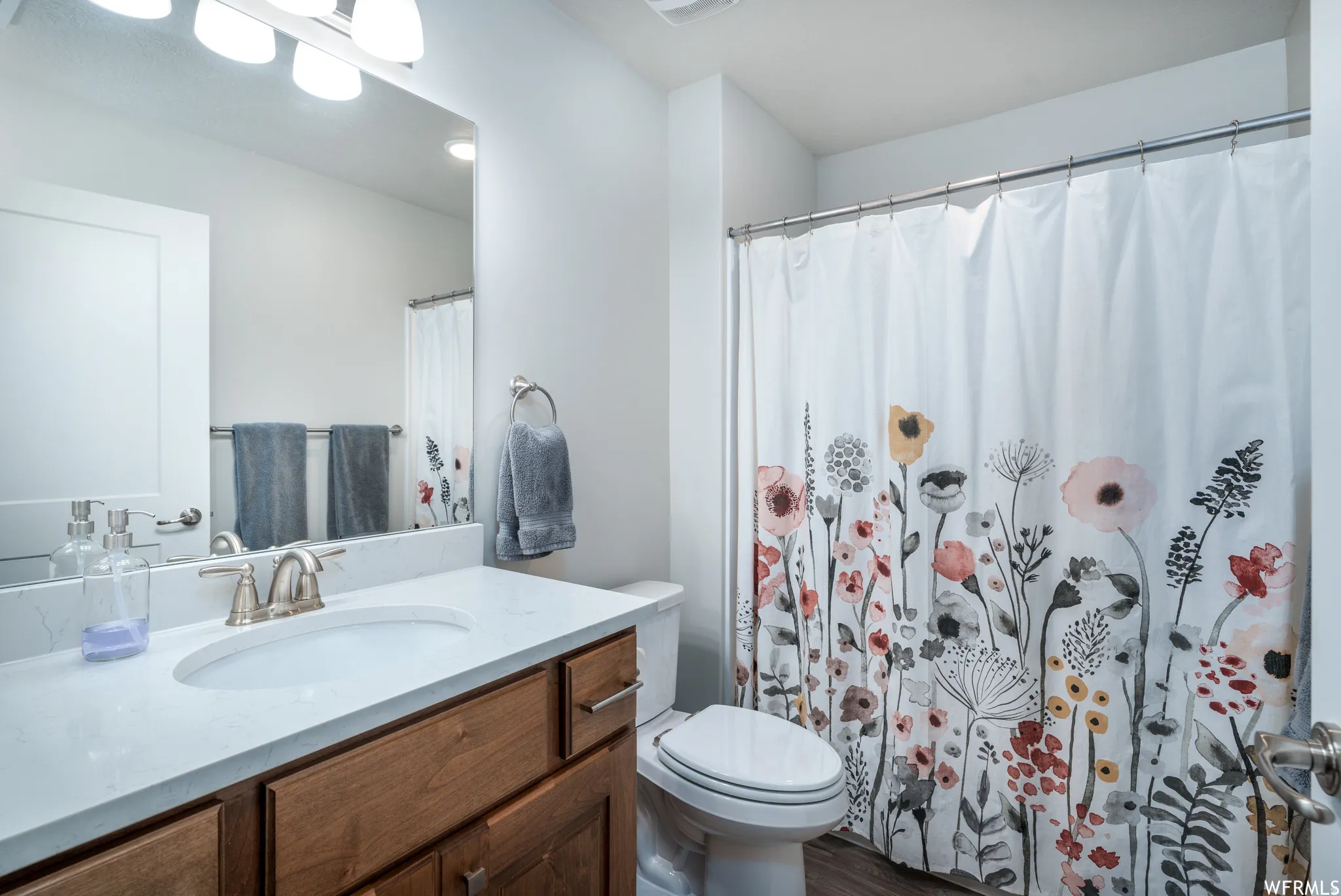 Bathroom featuring a vanity with a white countertop and an integrated sink, a wood-finish cabinet with brushed nickel hardware, and a framed mirror with a four-light fixture