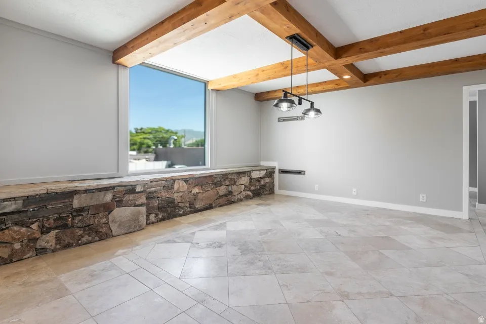 Spacious room featuring a large horizontal window, exposed wood ceiling beams, tiled flooring, a built-in stone ledge, and a contemporary linear pendant light fixture