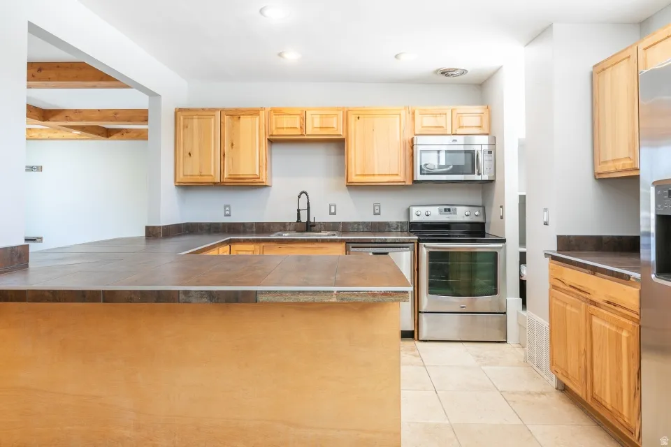 Kitchen featuring a U-shaped peninsula with tile countertops, light wood cabinetry, and stainless steel appliances including a range, dishwasher, microwave, and refrigerator