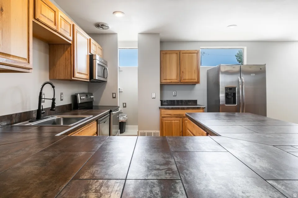 Kitchen featuring durable tile countertops, wood cabinetry, stainless steel appliances, and a modern black gooseneck faucet