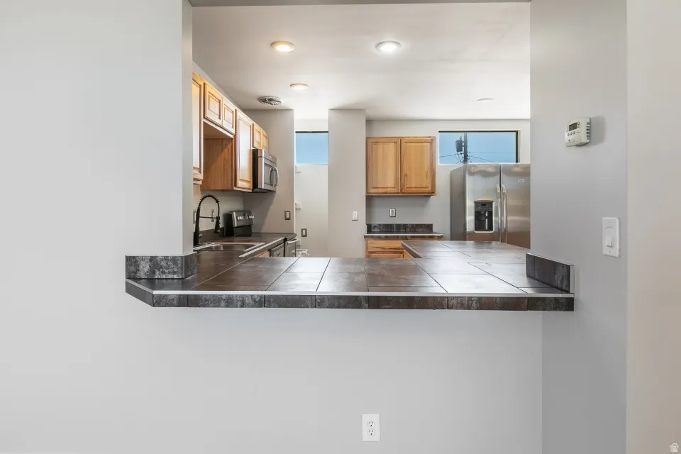 Kitchen pass-through featuring a tiled countertop, recessed lighting, wood cabinetry, stainless steel appliances, and a window