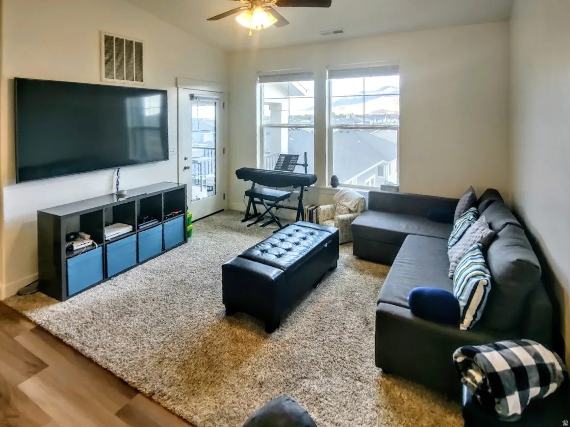 Living area featuring a textured carpet, wood-finish flooring, a ceiling fan, and abundant natural light from multiple windows and a glass-paneled door