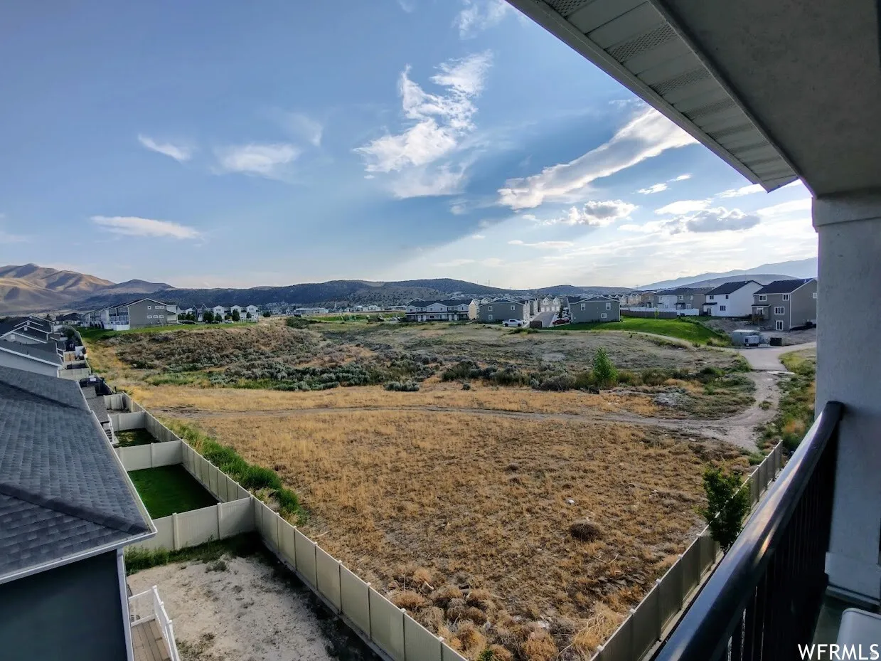 Expansive open land with distant mountain ranges, bordered by residential properties featuring light-colored fencing and a paved pathway