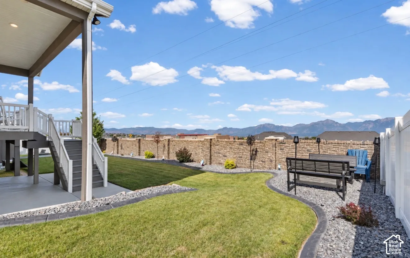 Expansive backyard featuring a manicured lawn, a covered concrete patio, a raised deck with stairs, a decorative stone retaining wall, and a white privacy fence