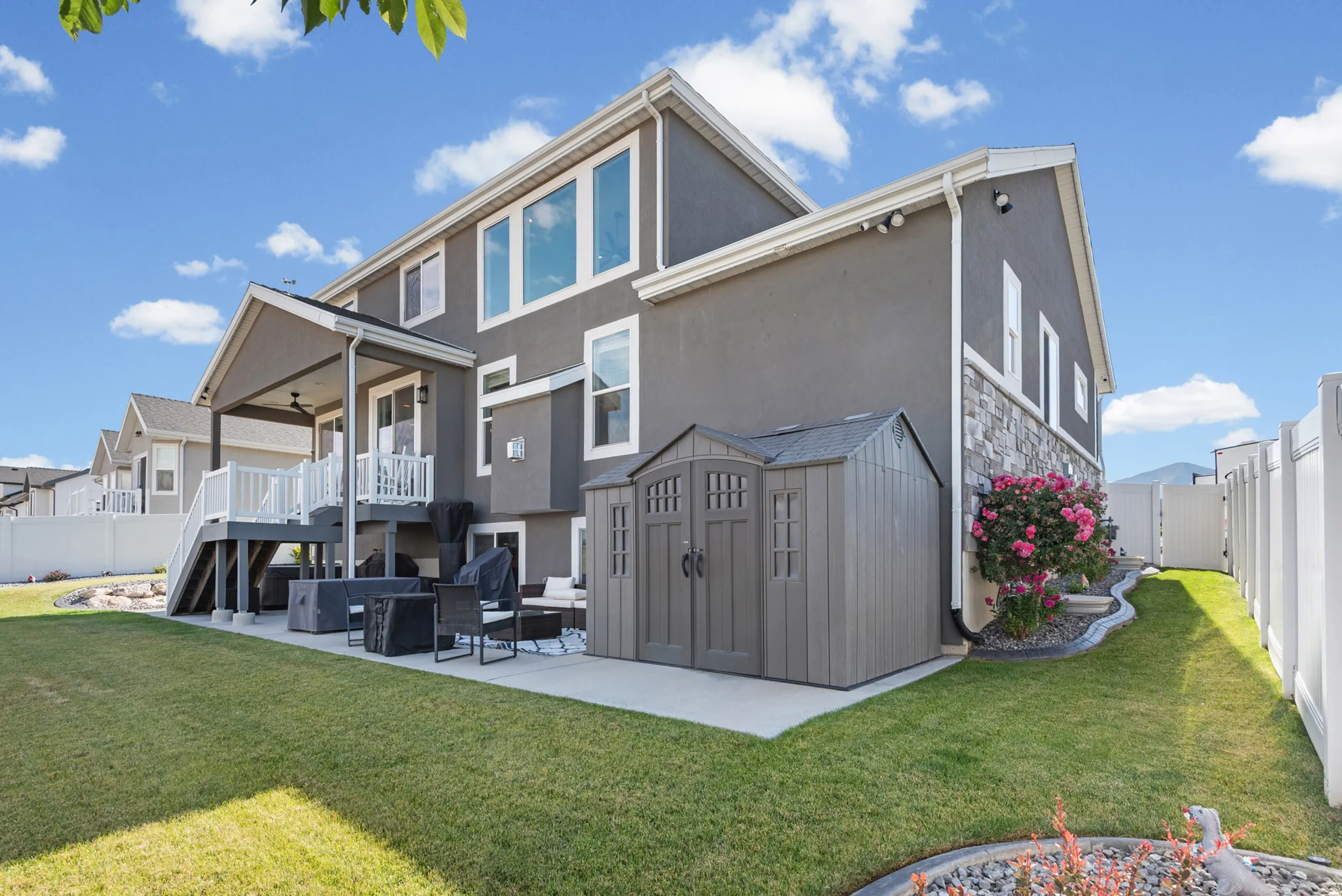 Rear exterior featuring a covered deck with white railings, a ground-level patio, a storage shed, and a stone facade accent