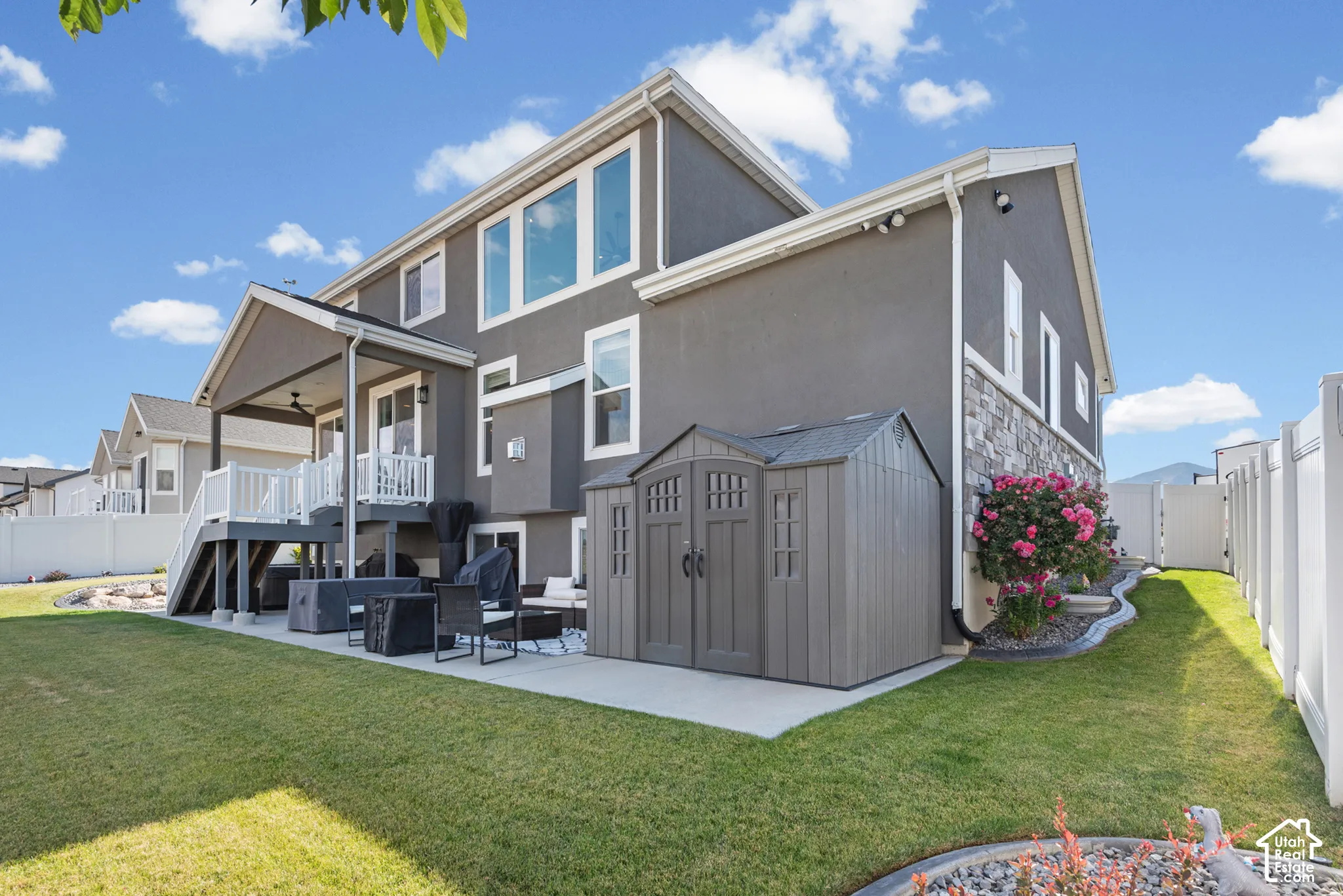 Rear exterior featuring a covered deck with white railings, a ground-level patio, a storage shed, and a stone facade accent