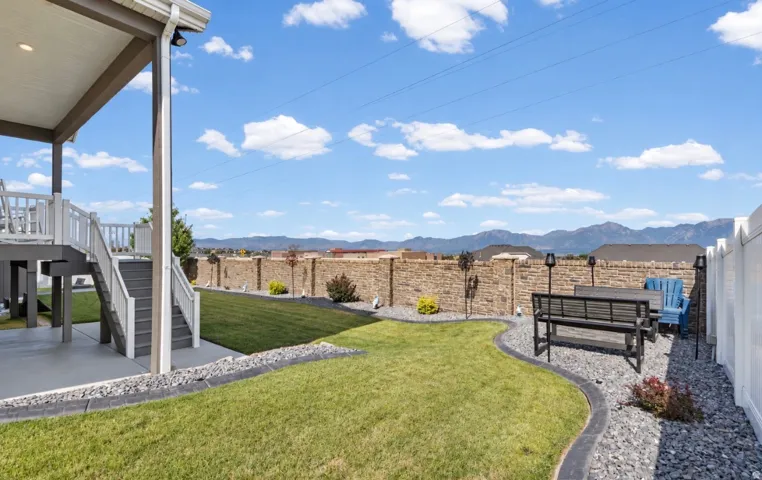 Expansive backyard featuring a manicured lawn, a covered concrete patio, a raised deck with stairs, a decorative stone retaining wall, and a white privacy fence