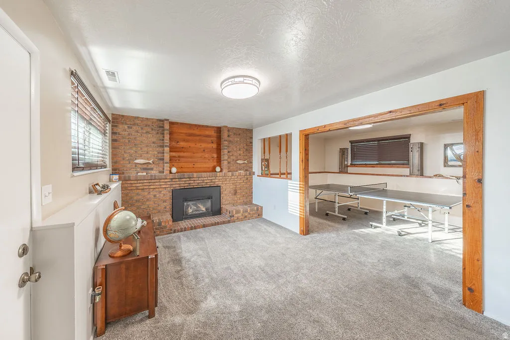 Brick fireplace with wood paneling accent, carpet flooring, a window with blinds, and an open doorway to an additional space