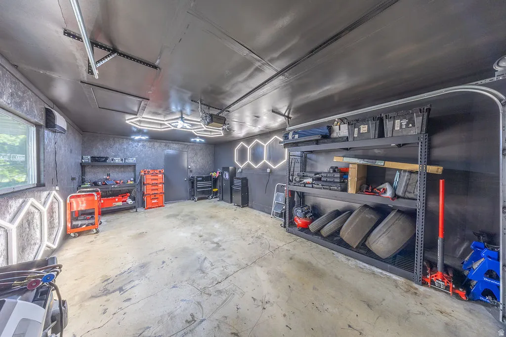 Spacious garage featuring concrete flooring, a window providing natural light, overhead and wall-mounted hexagonal lighting, extensive shelving for storage, and a ceiling-mounted HVAC unit