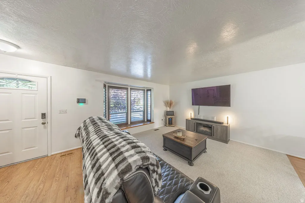 Spacious living area featuring light-toned carpeting, a prominent bay window with blinds, and a white entry door with decorative glass