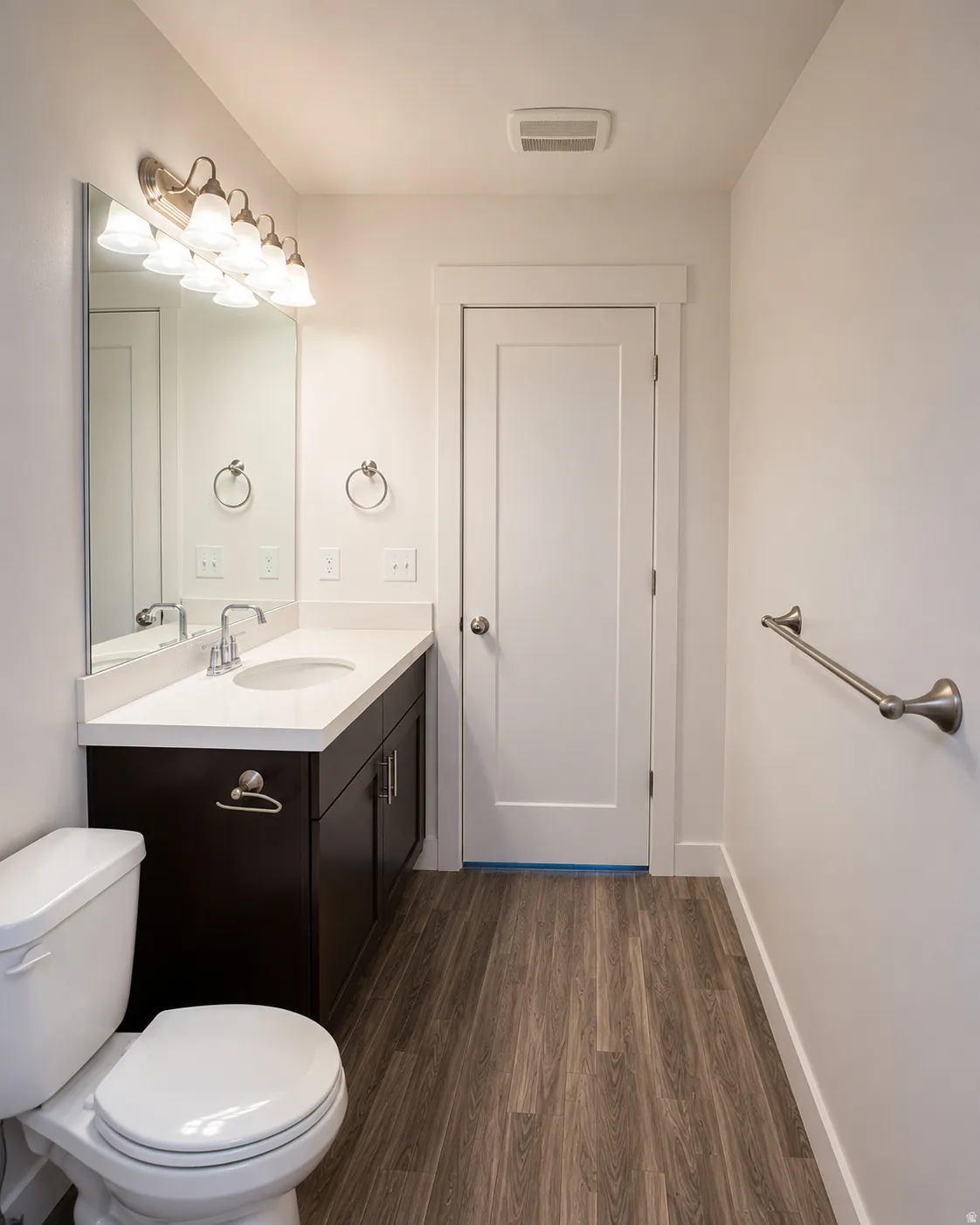 Bathroom featuring a dark wood-finish vanity with a white countertop and undermount sink, brushed nickel fixtures, wood-finish flooring, and a contemporary white door