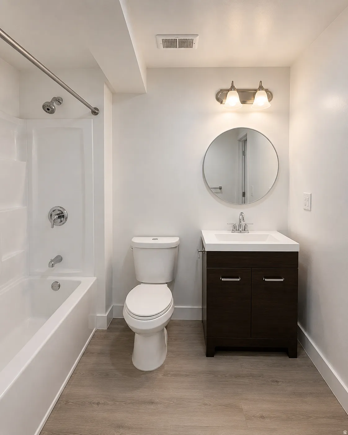 Bathroom featuring a white tub/shower combination, a dark wood-finish vanity with a white integrated sink, a round wall-mounted mirror, and wood-finish flooring