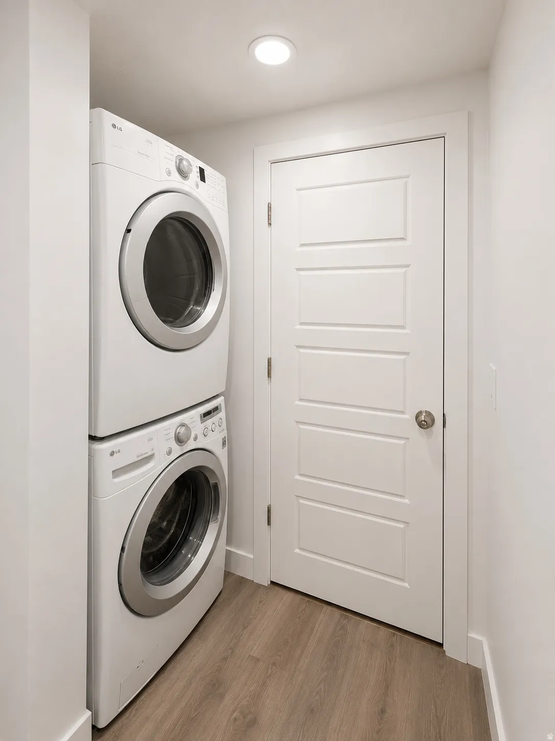 Dedicated laundry area featuring a stacked washer and dryer, wood-finish flooring, a contemporary paneled door, flush-mount lighting, and crisp white baseboards
