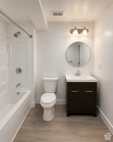 Bathroom featuring a white tub/shower combination, a dark wood-finish vanity with a white integrated sink, a round wall-mounted mirror, and wood-finish flooring