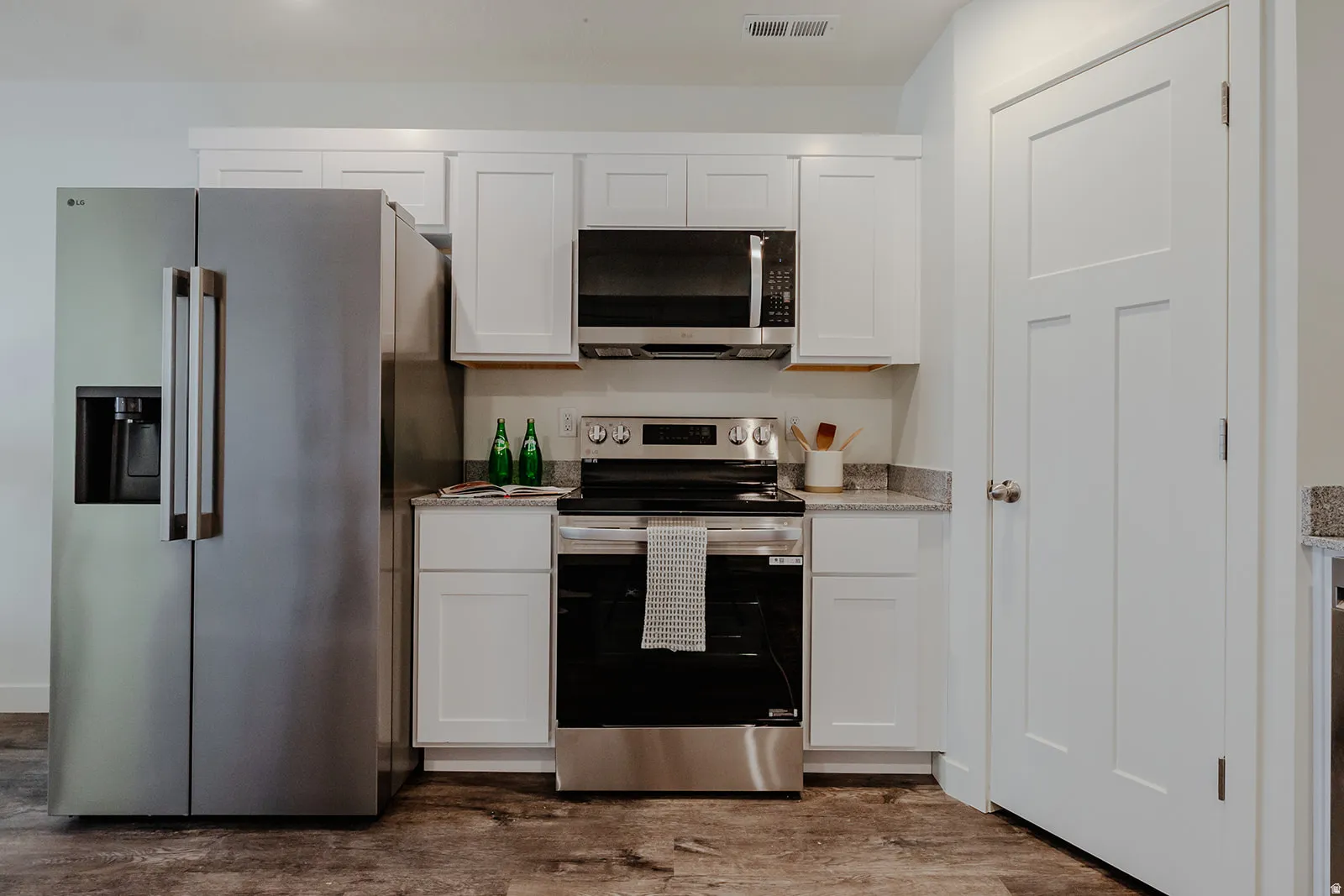 Modern kitchen featuring stainless steel appliances, white shaker cabinetry, granite-finish countertops, wood-finish flooring, and a contemporary panel door