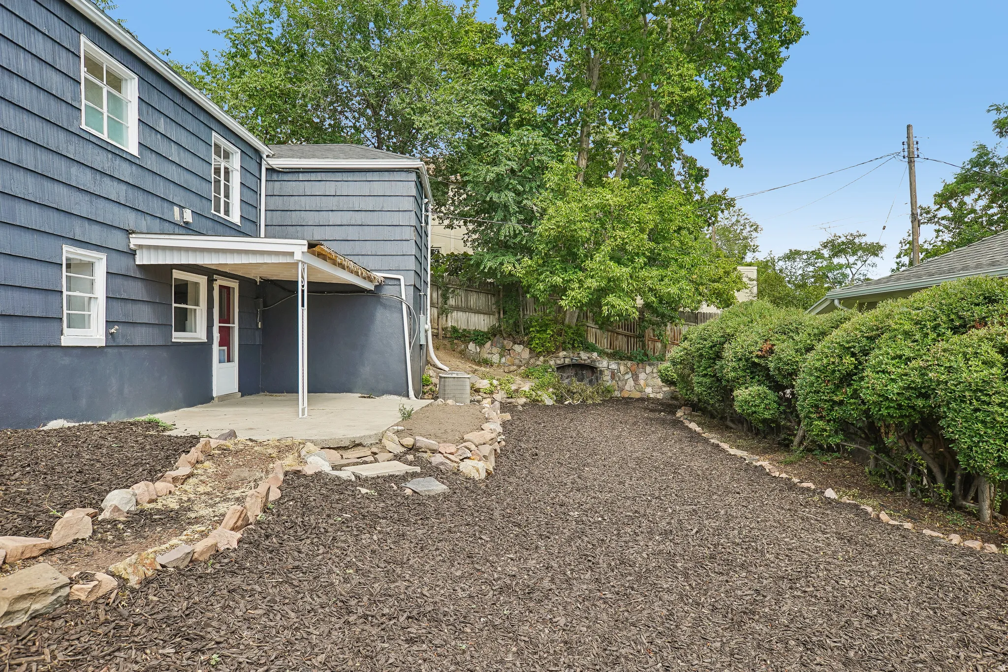 Rear exterior featuring a covered concrete patio, wood chip landscaping, stone retaining walls, and mature perimeter trees