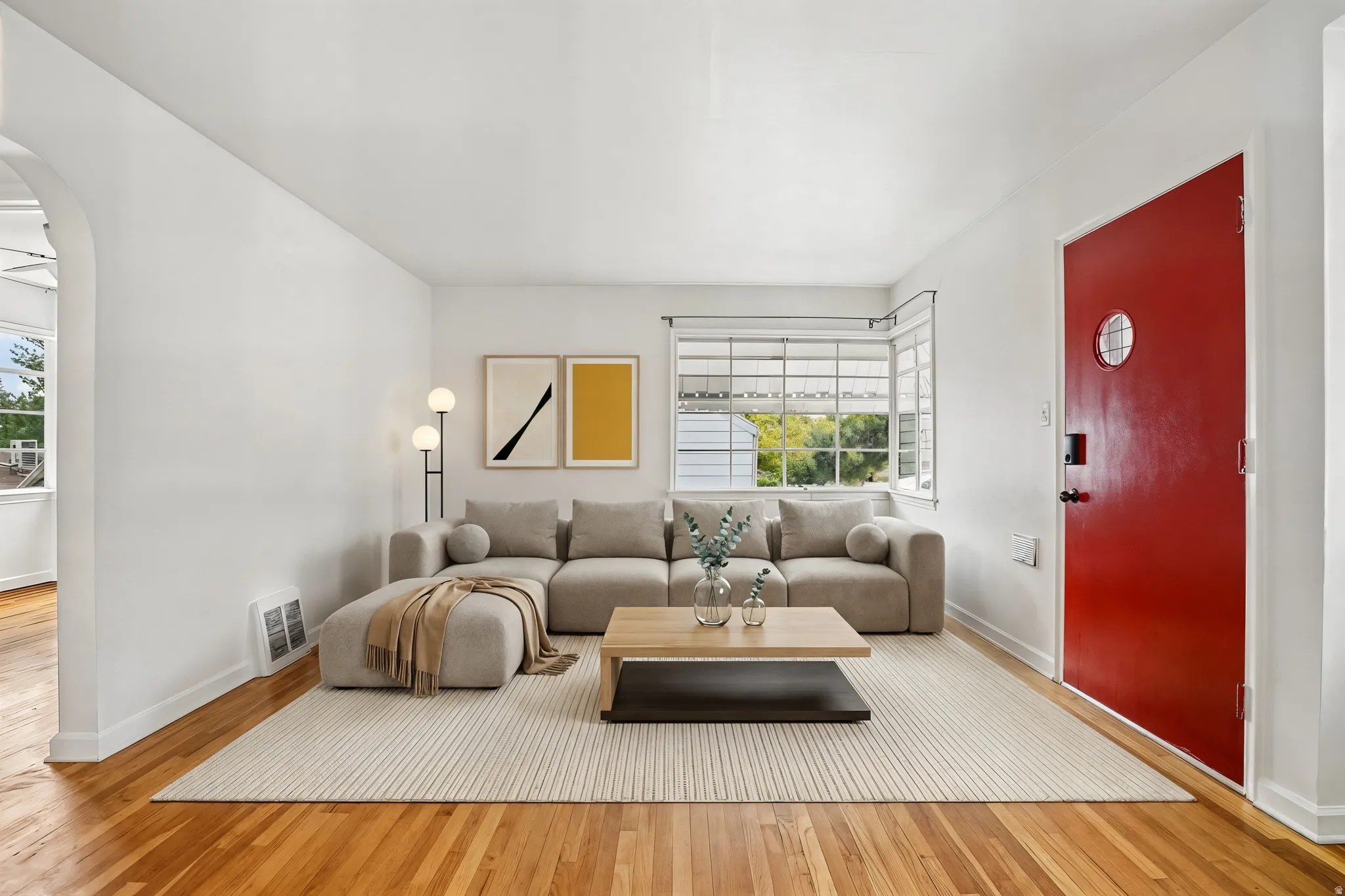 Bright living area featuring wood-finish flooring, white walls, and a prominent red entry door with a circular window