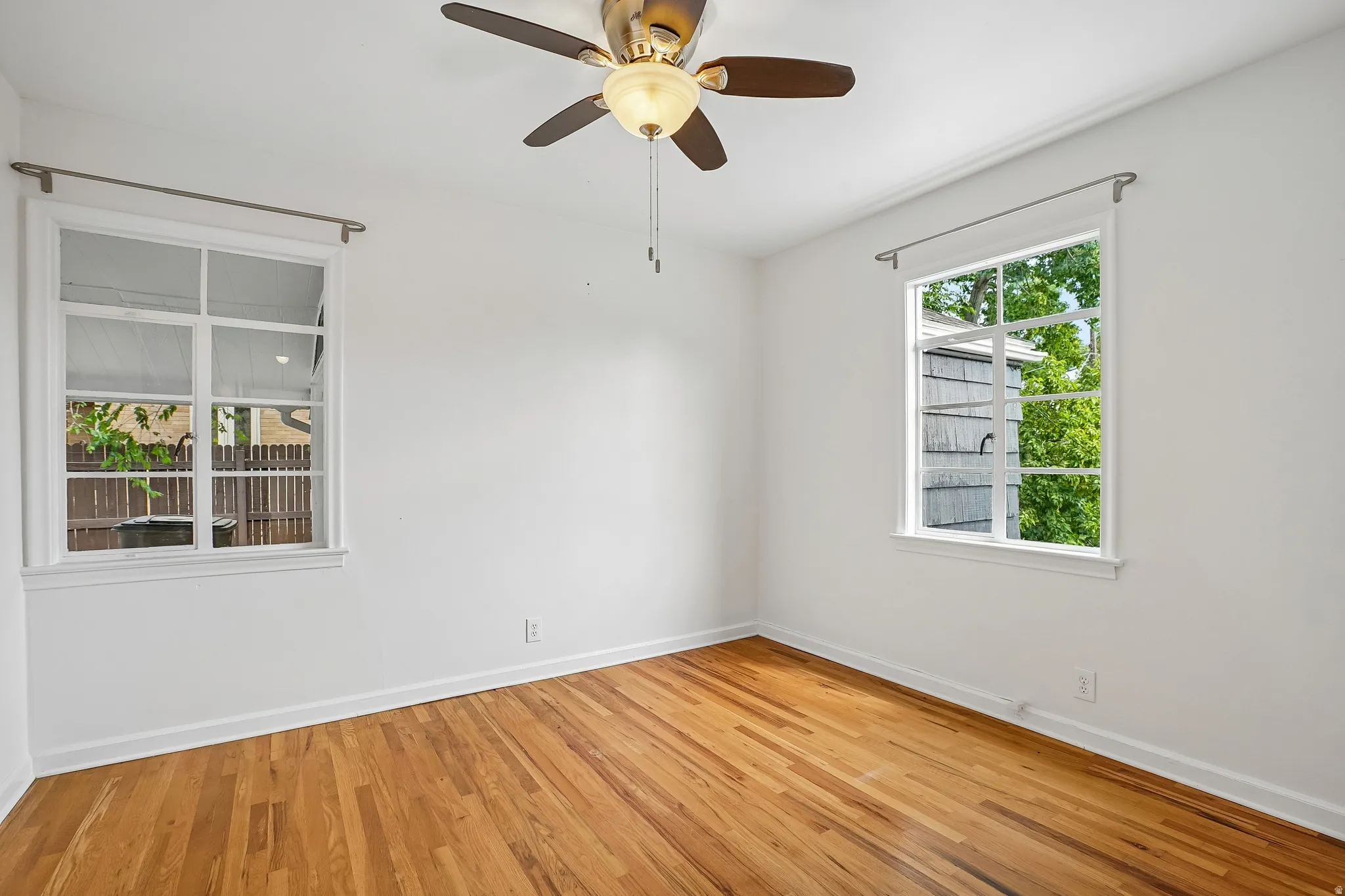 Hardwood flooring complemented by white baseboards, two windows with white trim, and a ceiling fan with integrated lighting