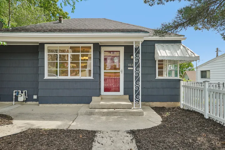 Exterior featuring dark gray horizontal siding, a red entry door, a white decorative porch column, a white picket fence, and a window with a white awning
