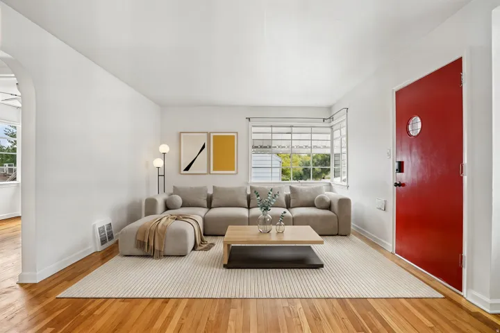 Bright living area featuring wood-finish flooring, white walls, and a prominent red entry door with a circular window