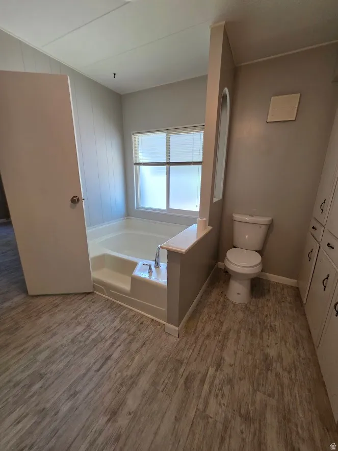Bathroom featuring a built-in tub with chrome fixtures, a privacy window, a commode, and wood-finish flooring