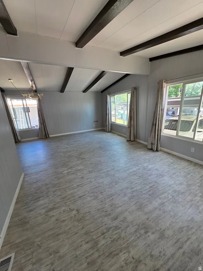 Spacious interior featuring a vaulted ceiling with exposed dark-toned beams, light gray wood-finish flooring, light gray paneled walls, and multiple windows with valance and drapes