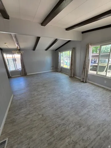 Spacious interior featuring a vaulted ceiling with exposed dark-toned beams, light gray wood-finish flooring, light gray paneled walls, and multiple windows with valance and drapes