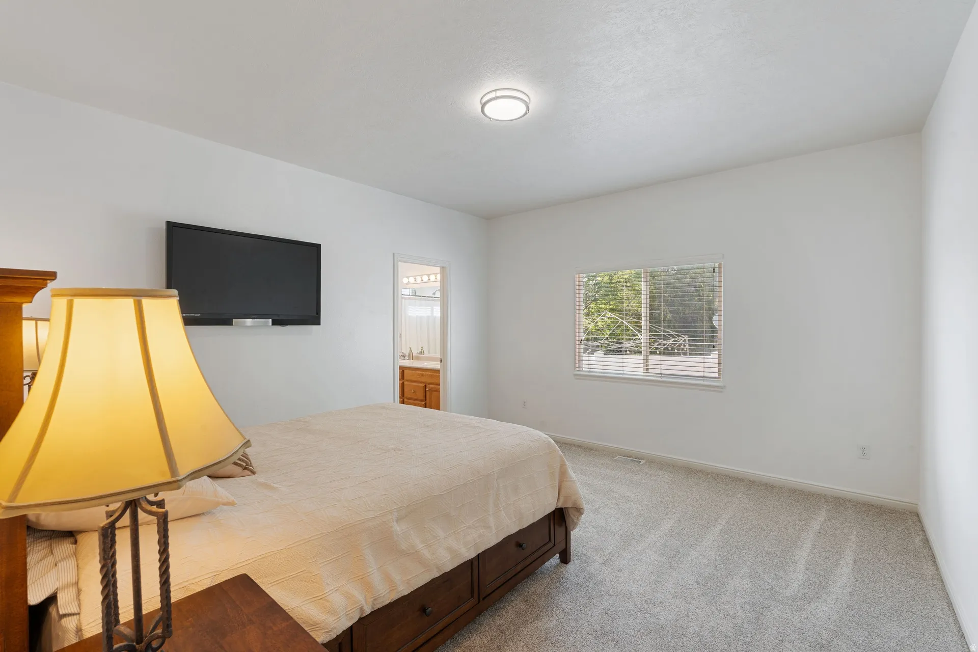 Bright room featuring a window with blinds, light-toned carpet, a ceiling-mounted light fixture, and a doorway leading to an adjacent space with a wood-finish vanity