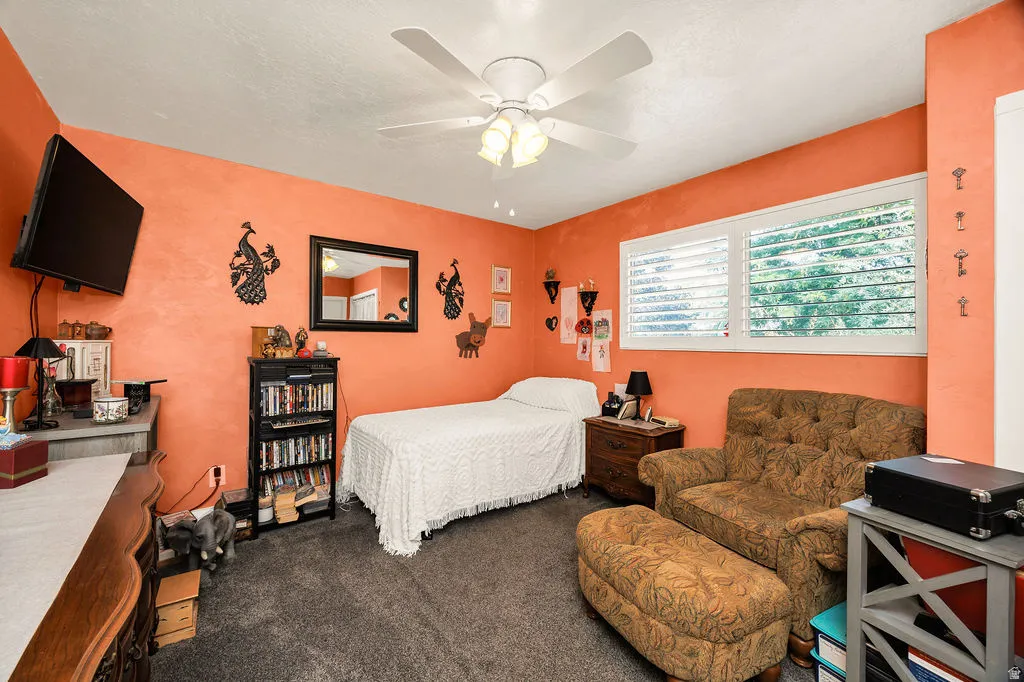 Interior room featuring textured orange walls, gray carpet, a white ceiling fan with integrated lighting, and a large window with white plantation shutters